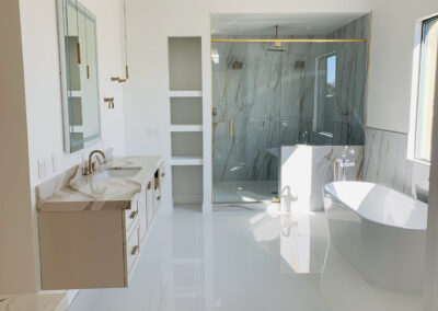 A powder room designed and built by Armstrong Construction Group in Arizona. Features a floating vanity with a marble counter to, glossy white flooring, a white tub under a large window, and a glass walled shower with marbled tiles.