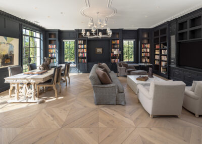 A home library built by Armstrong Construction Group with dark walls covered with books and shelving, a unique patterned wood floor, light colored table and lounge furniture. The room has ample natural light flowing in from the windows around the room.