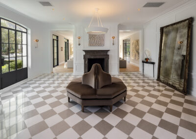 A foyer designed and built by Armstrong Construction Group. Features checkered flooring, a large mirror, large windows to the left, two doorways on either side of a fireplace and a settee in the middle with a hanging lamp above it.