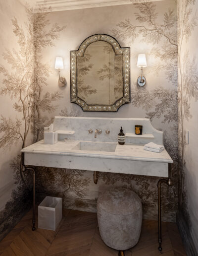 A custom luxury bathroom designed by Armstrong Construction Group. Features a white marble vanity, an ornate mirror with sconce lights on both sides. The walls have a cream and brown wall paper with a tree design. The floor is a lasercut wood floor running throughout the home.