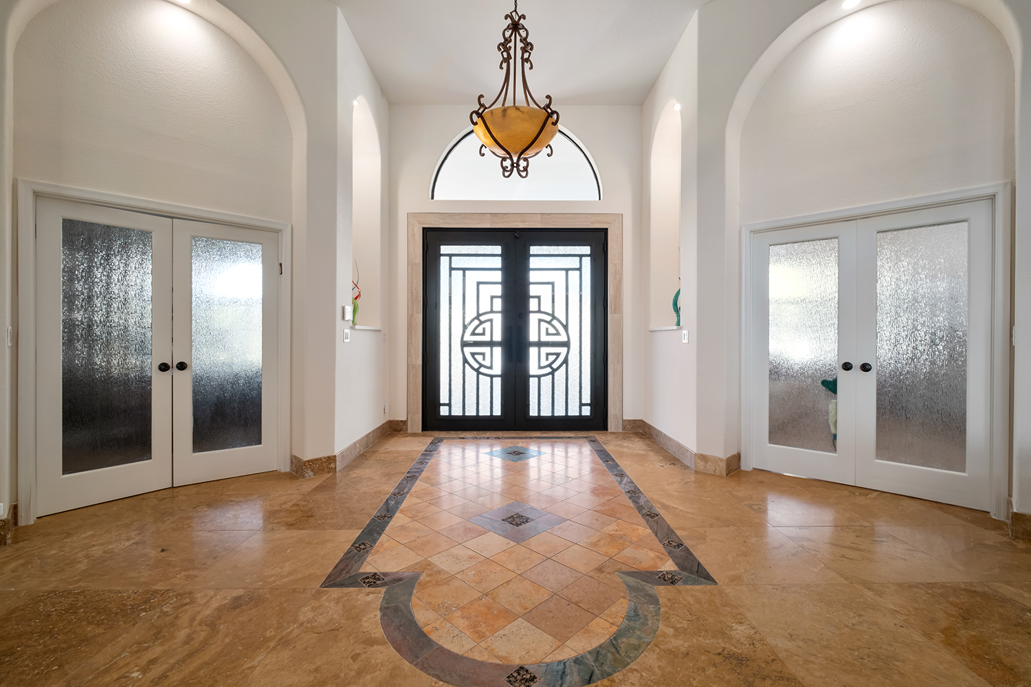 A home entryway designed by Armstrong Construction Group in Arizona. Features a large black angular door, tan marble flooring, and doors on either side of the entryway leading into new rooms.