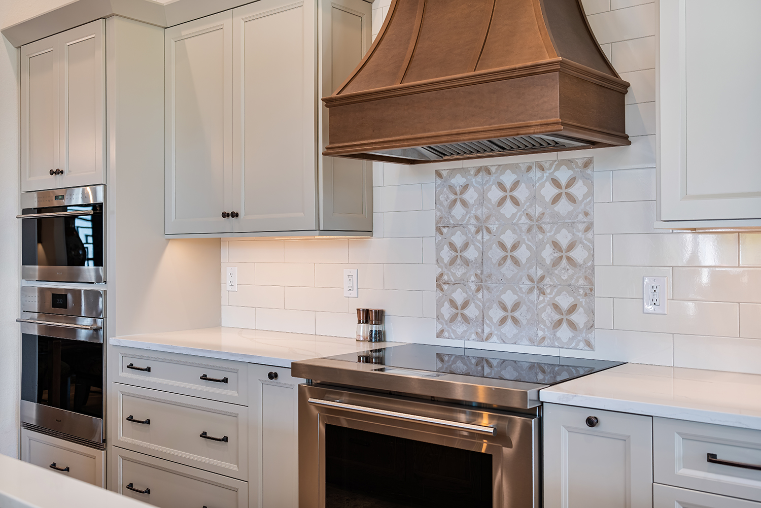 Custom built kitchen by Armstrong Construction Group. Shows kitchen oven and stove with bronze vent, light grey cabinets, white countertops and an ornate tile backsplash.
